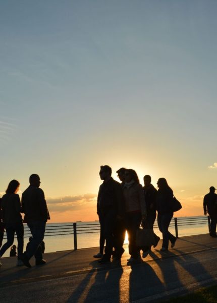 Silhouettes of people walking along a waterfront during a vibrant sunset casting long shadows.